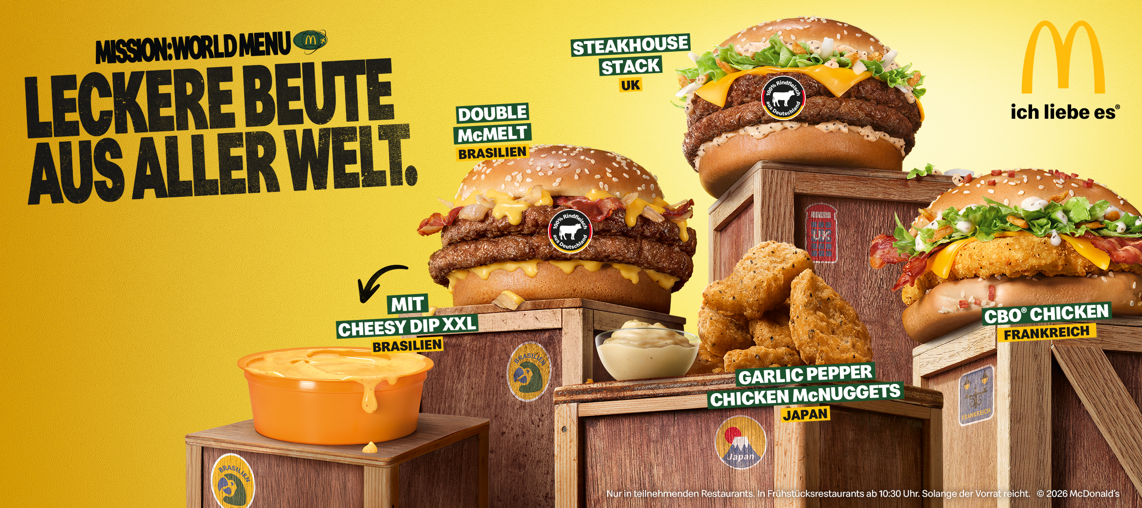 Werbebanner von McDonald's zur Aktion „Mission: World Menu“. Vor gelbem Hintergrund sind fünf Aktionsprodukte auf Holzkisten zu sehen: „Double McMelt (Brasilien)“, „Steakhouse Stack (UK)“, „CBO Chicken (Frankreich)“, „Garlic Pepper Chicken McNuggets (Japan)“ und „Cheesy Dip XXL (Brasilien)“. Links steht der Slogan „MISSION: WORLD MENU – LECKERE BEUTE AUS ALLER WELT.“, rechts das McDonald's-Logo mit dem Schriftzug „ich liebe es®“.
