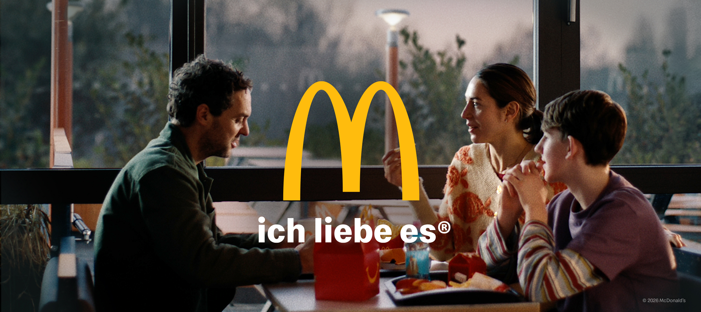 Vater, Mutter, Sohn sitzen an einem Tisch in einem McDonald’s Restaurant, essen Produkte eines Happy Meal's® und unterhalten sich in aufgelockerter Stimmung. Im Vordergrund das gelbe McDonald’s "M", darunter der Slogan "ich liebe es®"