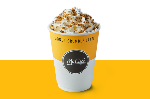 A McCafé Donut Crumble Latte topped with whipped cream and caramelised crumble pieces, served in a yellow and white takeaway cup.