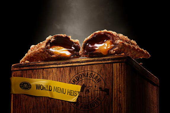 McDonald’s Choco Caramle Pie with goeey chocolate and caramel filling, shown halved on a wooden crate with 'World Menu Heist' tape, featured against dark background.