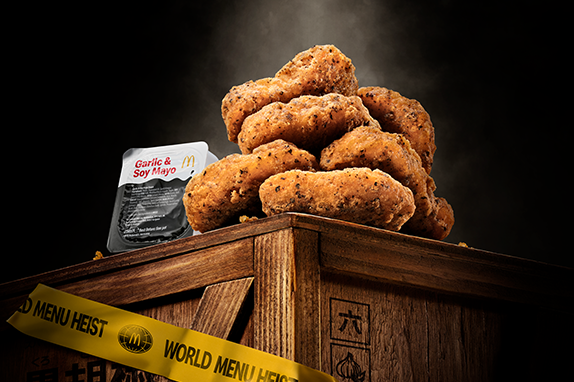 McDonald’s Black Pepper & Garlic Chicken McNuggets® with Garlic & Soy May sauce dip packet, displayed on a wooden crate with 'World Menu Heist' tape, featured against dark background.