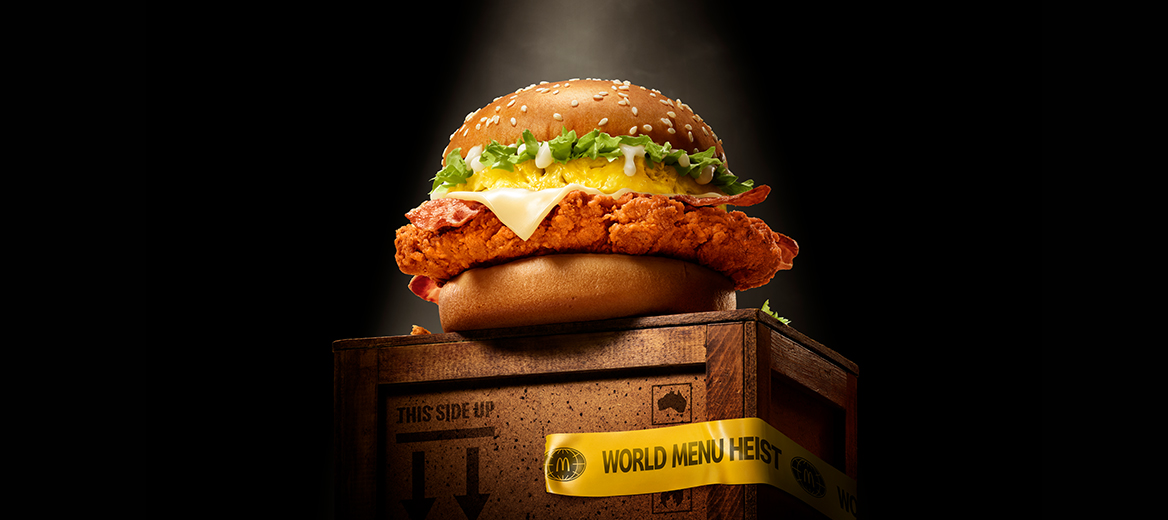A McDonald’s Pineapple McSpicy® burger displayed on a wooden crate with 'World Menu Heist' tape, featured against dark background.