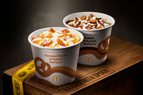 Two McDonald’s Pretzel McFlurry cips, Salted Caramel and Chocolate, displayed on a wooden crate with 'World Menu Heist' tape, featured against dark background.