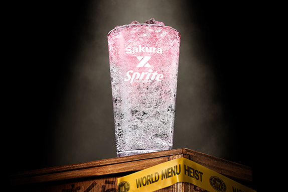A McDonald’s glass with a mix of Sprite Zero and sugar-free Sakura Cherry Blossom flavoured syrup, served over ice, displayed a wooden crate with 'World Menu Heist' tape, featured against dark background.