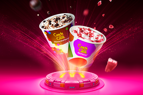 Candy Crush Choco McFlurry floating above a glowing McDonald's platform, with chocolate-coated candy pieces suspended around the cup against a bright pink and purple background.