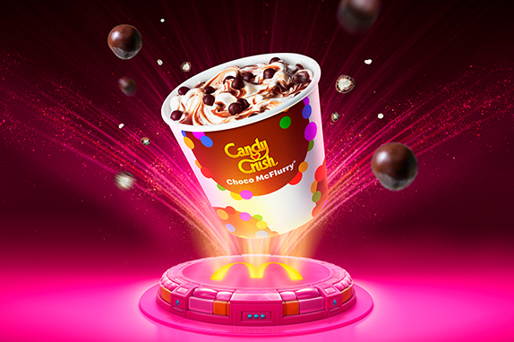 Candy Crush Choco McFlurry floating above a glowing McDonald's platform, with chocolate-coated candy pieces suspended around the cup against a bright pink and purple background.