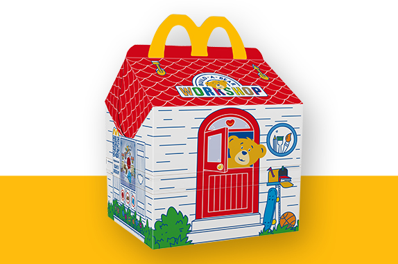 Red and white Build-A-Bear Happy Meal box with the bear peeping out of the window of the box on a yellow shelf 