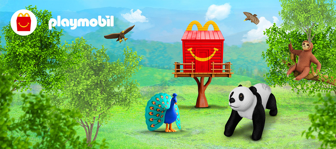 A red Happy Meal box treehouse sits in a field, with mountains in the backdrop and trees in the foreground. Playmobil eagles fly in the blue sky, a Playmobil sloth sits in a tree and a peacock and panda stand in front of the treehouse on the grass. 